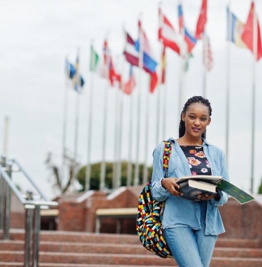 African student female posed with backpack and school items on yard of university, against flags of different countries.